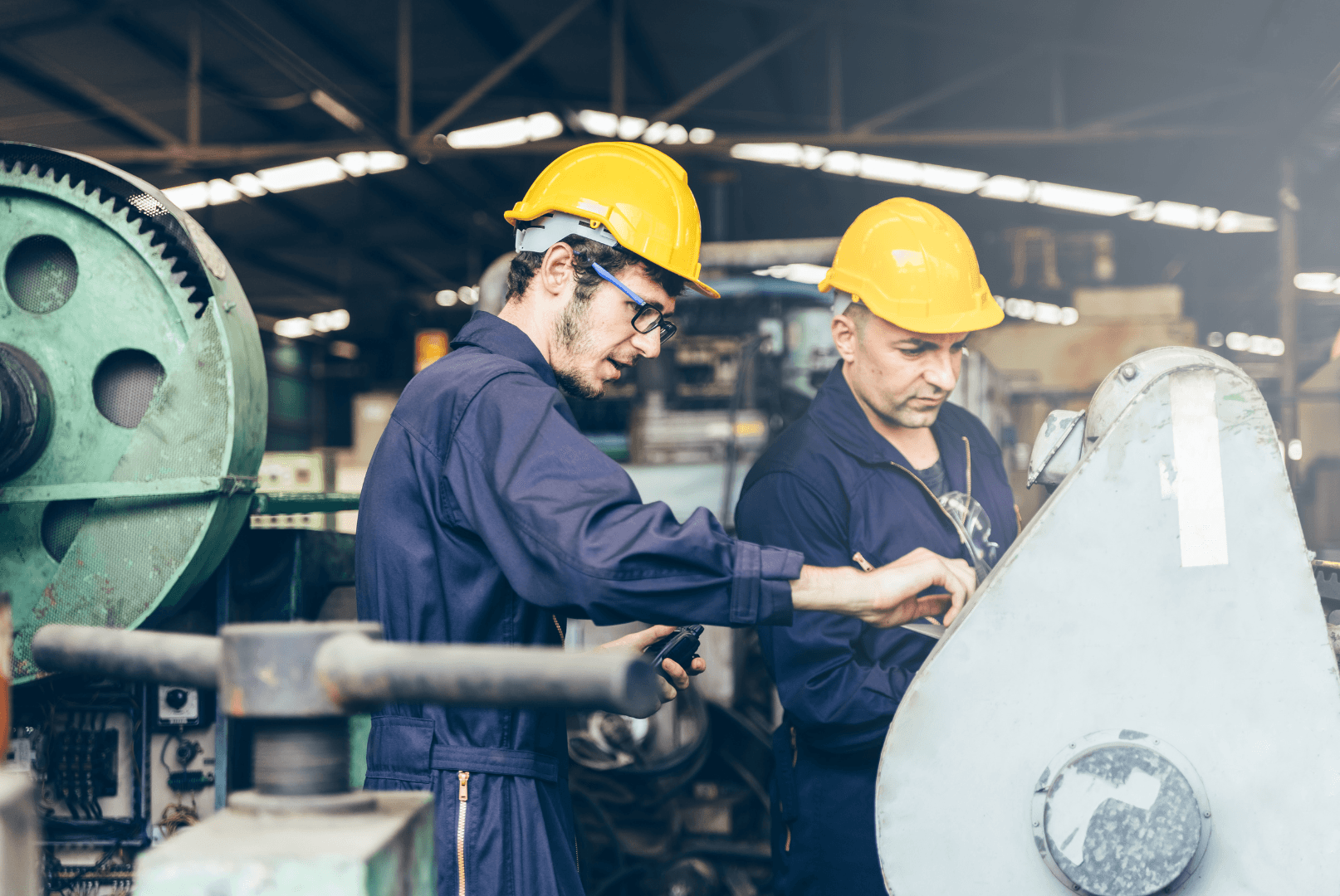 Dois técnicos de capacete e macacão examinando uma máquina em galpão industrial