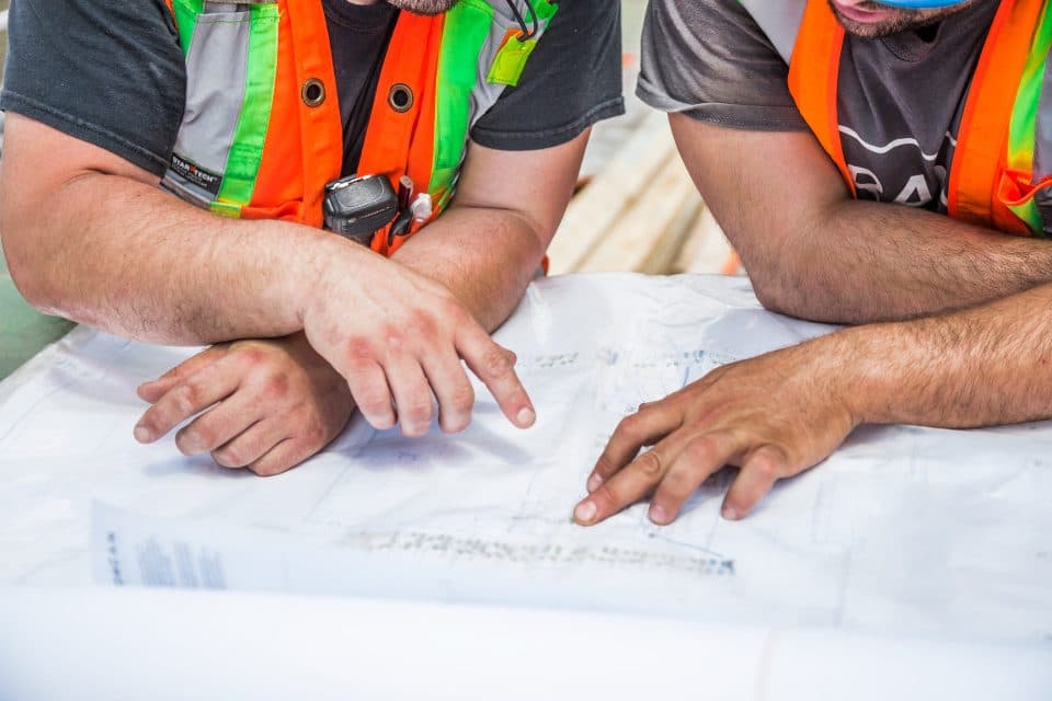 Duas pessoas de colete refletivo debruçadas sobre um desenho técnico, apontando um trecho