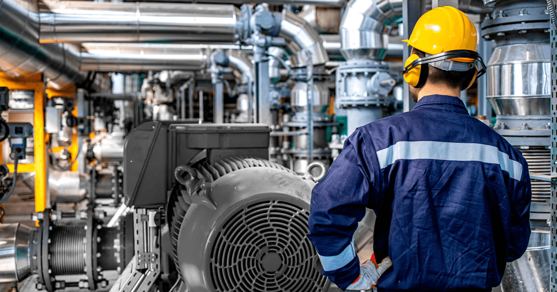 Técnico de capacete e protetor auricular de costas, diante de um motor elétrico em sala de tubulações